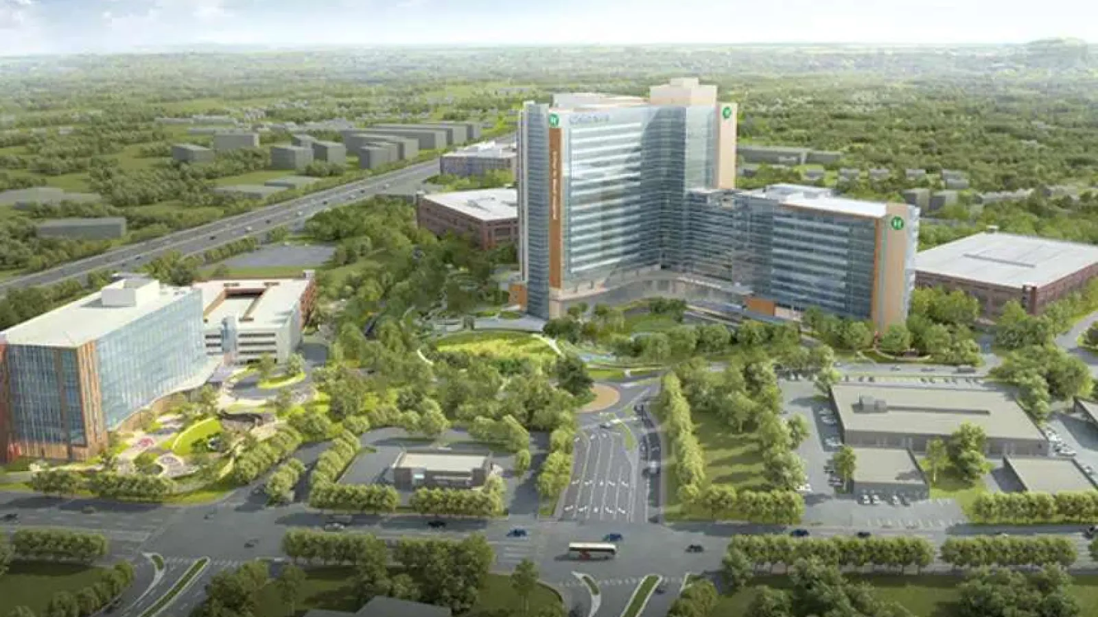 Aerial view of a modern hospital campus with multiple buildings surrounded by trees and roads in a green landscape.