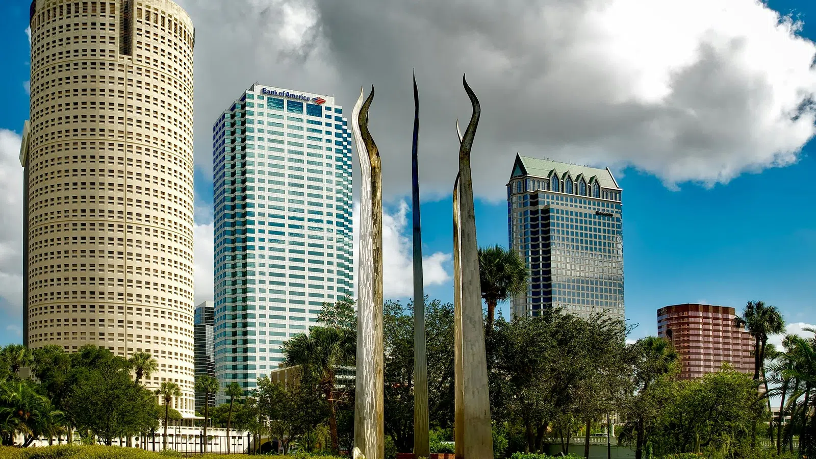 Tall modern skyscrapers and a unique metallic sculpture under a blue sky with clouds in a city park landscape.