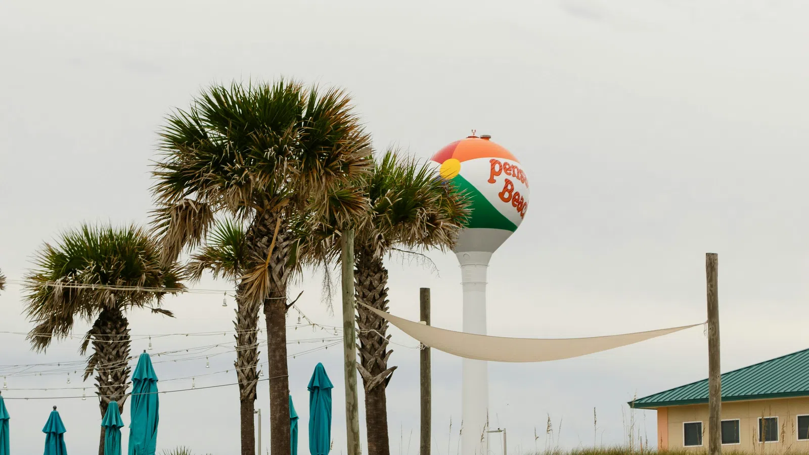 Empty blue lounge chairs on sandy beach with palm trees and a colorful Pensacola Beach water tower in the background.