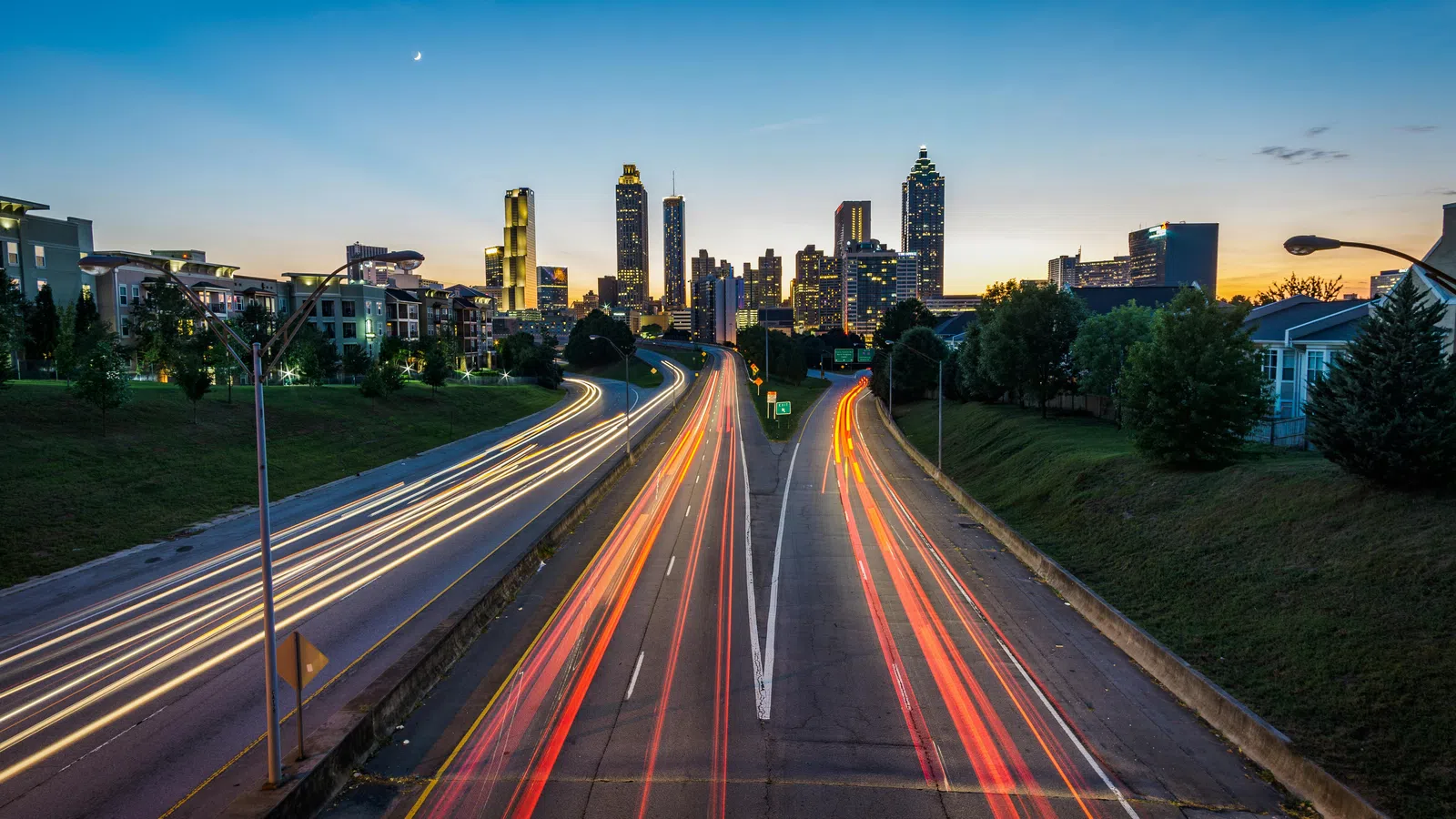 City skyline at dusk with light trails on a highway leading into downtown and a clear evening sky.