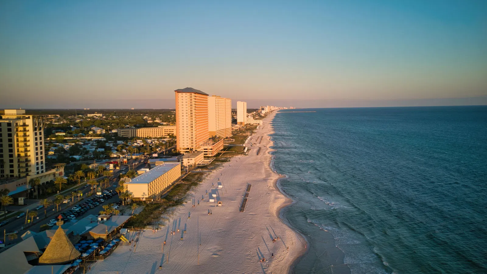 Aerial view of a coastal city with high-rise buildings along a sandy beach and calm ocean waters at sunset.