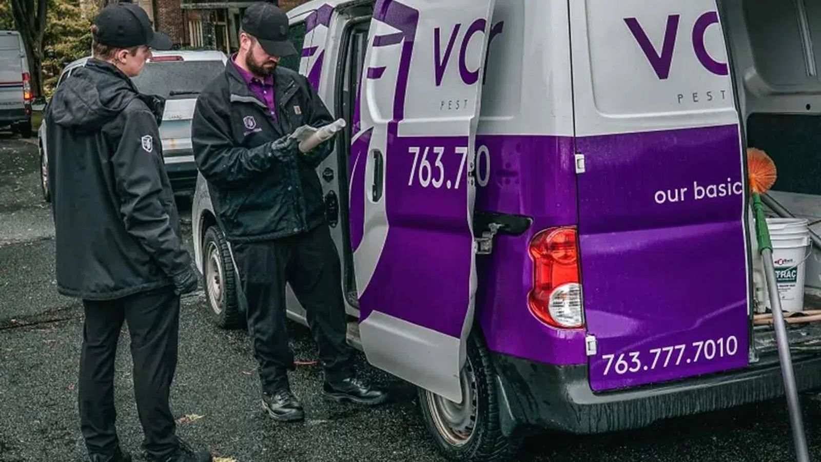 Two pest control workers in uniform consulting near a white and purple service van with equipment inside.