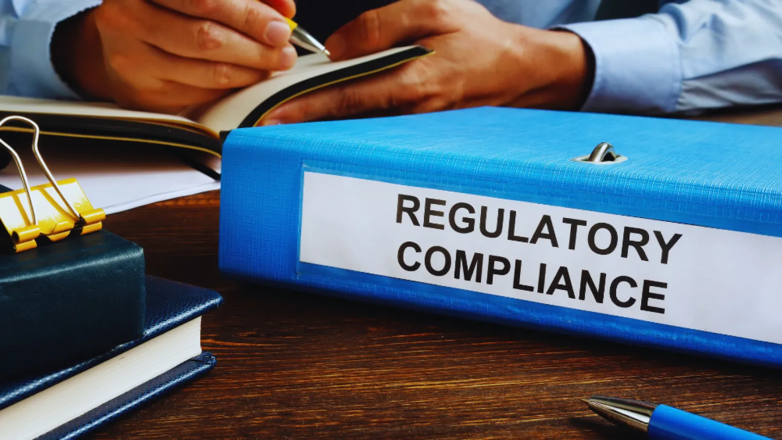 Person writing in notebook with blue binder labeled Regulatory Compliance on wooden desk.