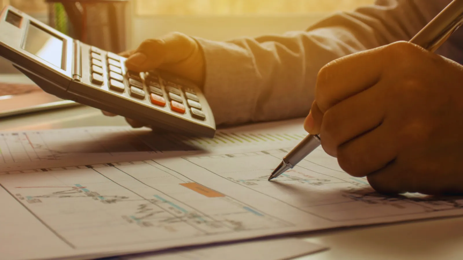Person using calculator and pen to analyze financial documents at a sunlit workspace.