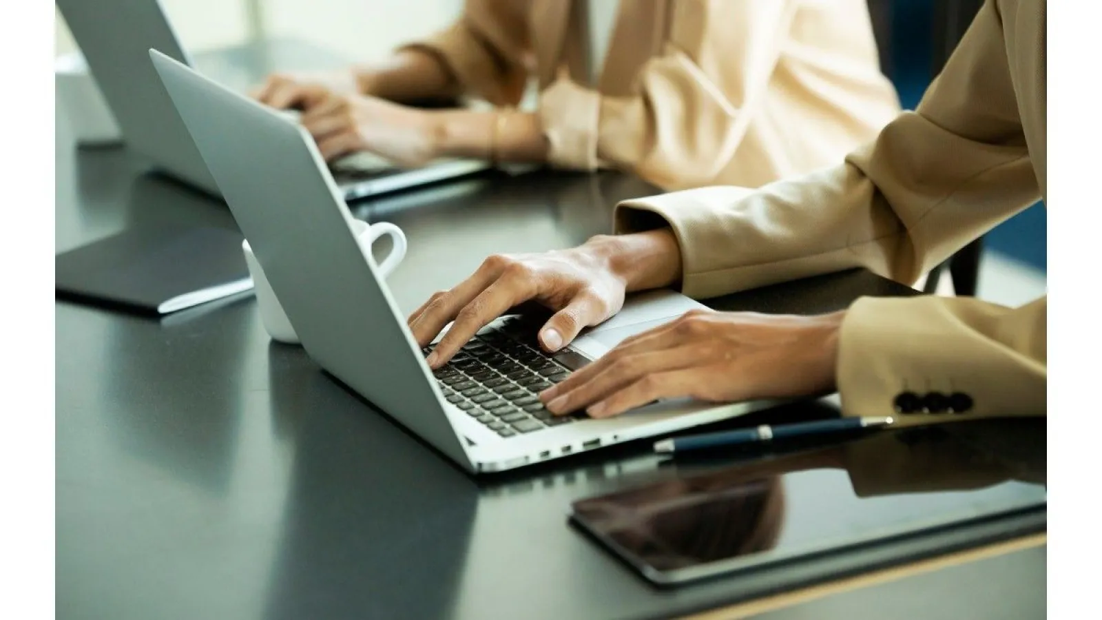 Two professionals typing on laptops at a desk with a coffee cup, notebook, and tablet nearby in an office setting