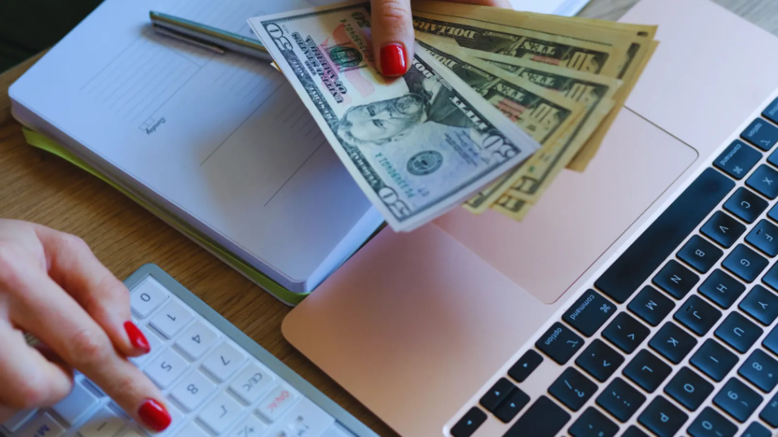 Person holding US dollar bills and using calculator with laptop and notebook on wooden desk