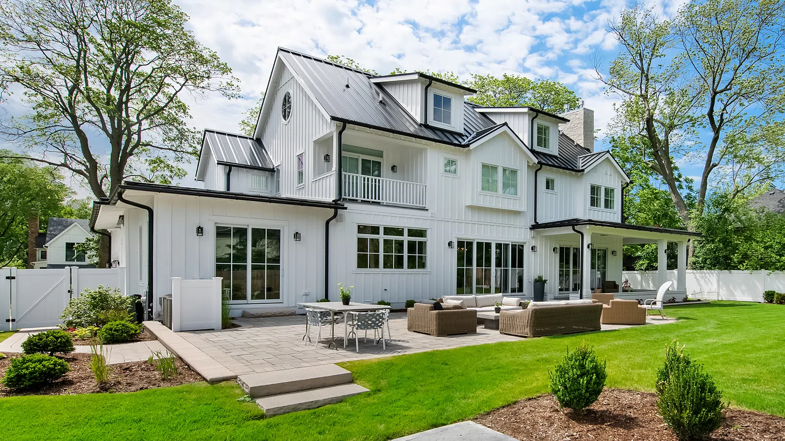 Modern white farmhouse with large windows, patio furniture, green lawn, and surrounding trees under blue sky.