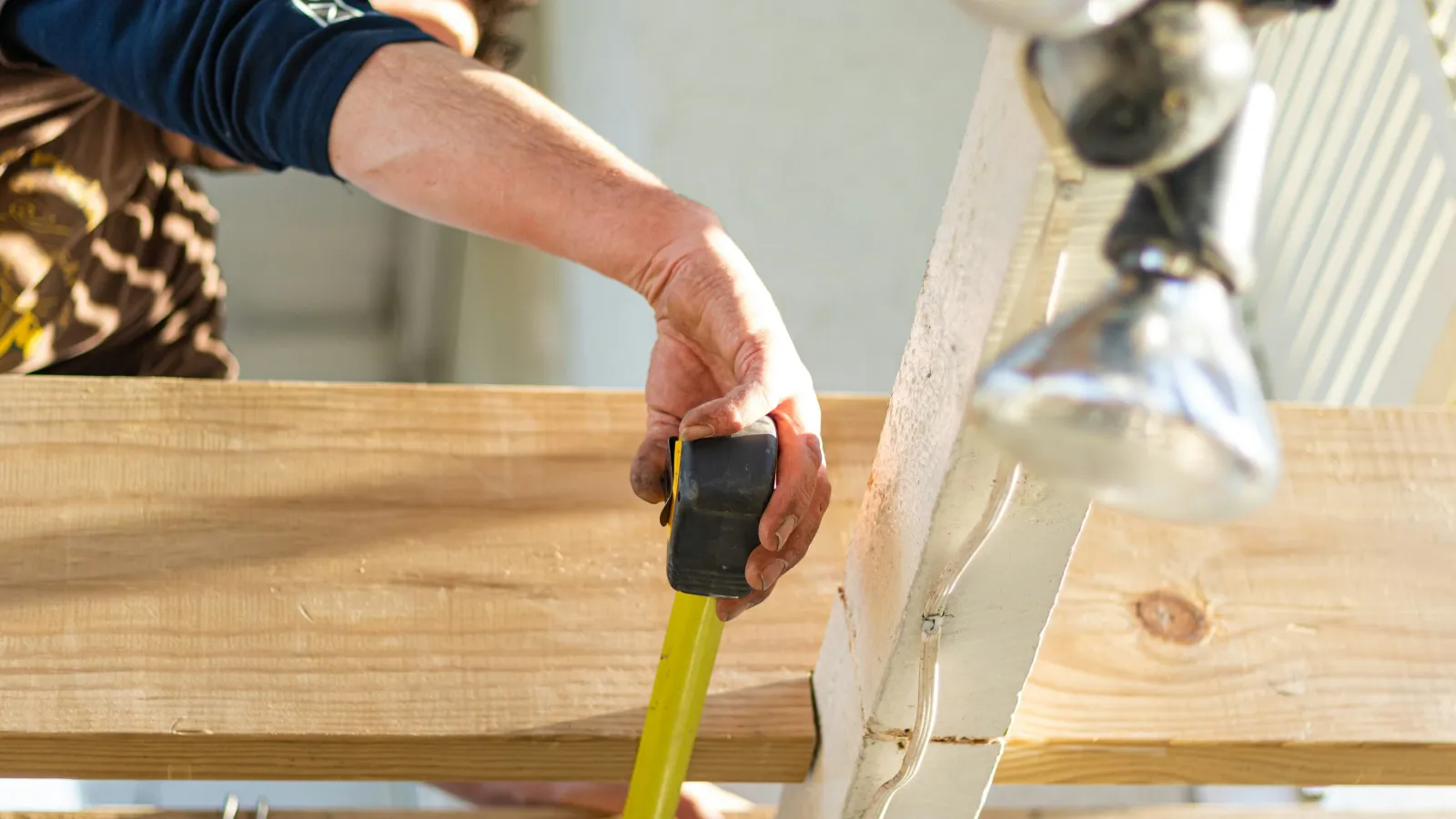 Person using a yellow tape measure to measure a wooden plank in a woodworking or construction setting.