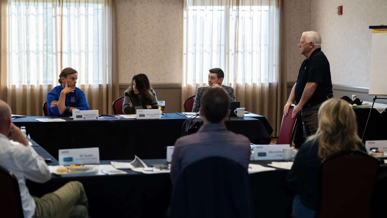 Business meeting with six professionals seated around tables discussing in a conference room with natural light.