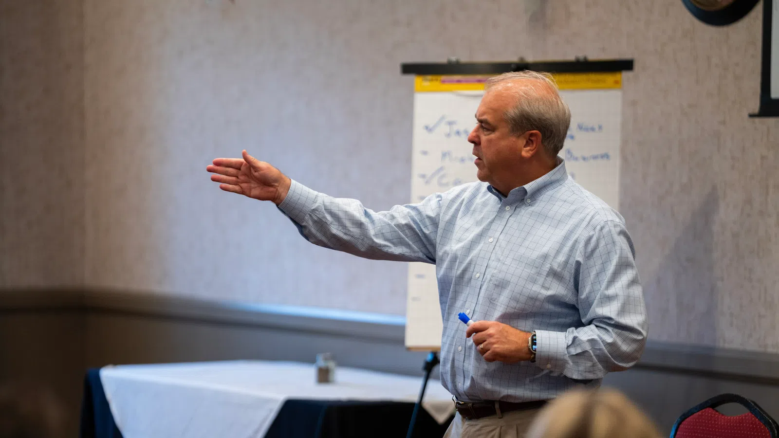 Mature man in button-up shirt speaking and gesturing during a meeting with flipchart and seated audience.