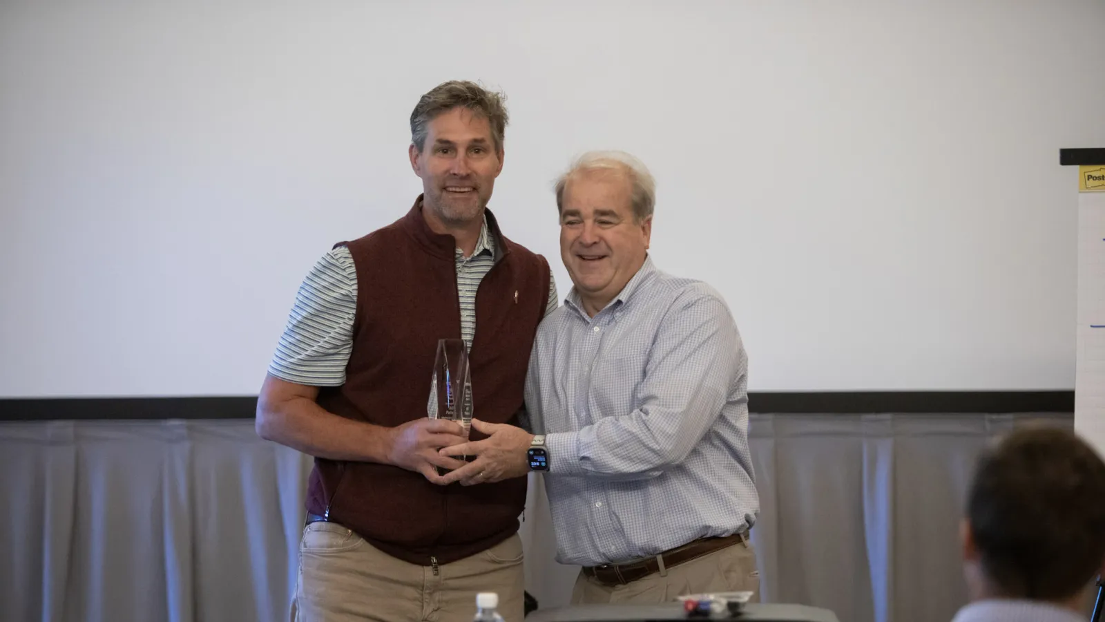 Two men smiling and holding a clear award trophy in a meeting room with a projector and attendees.