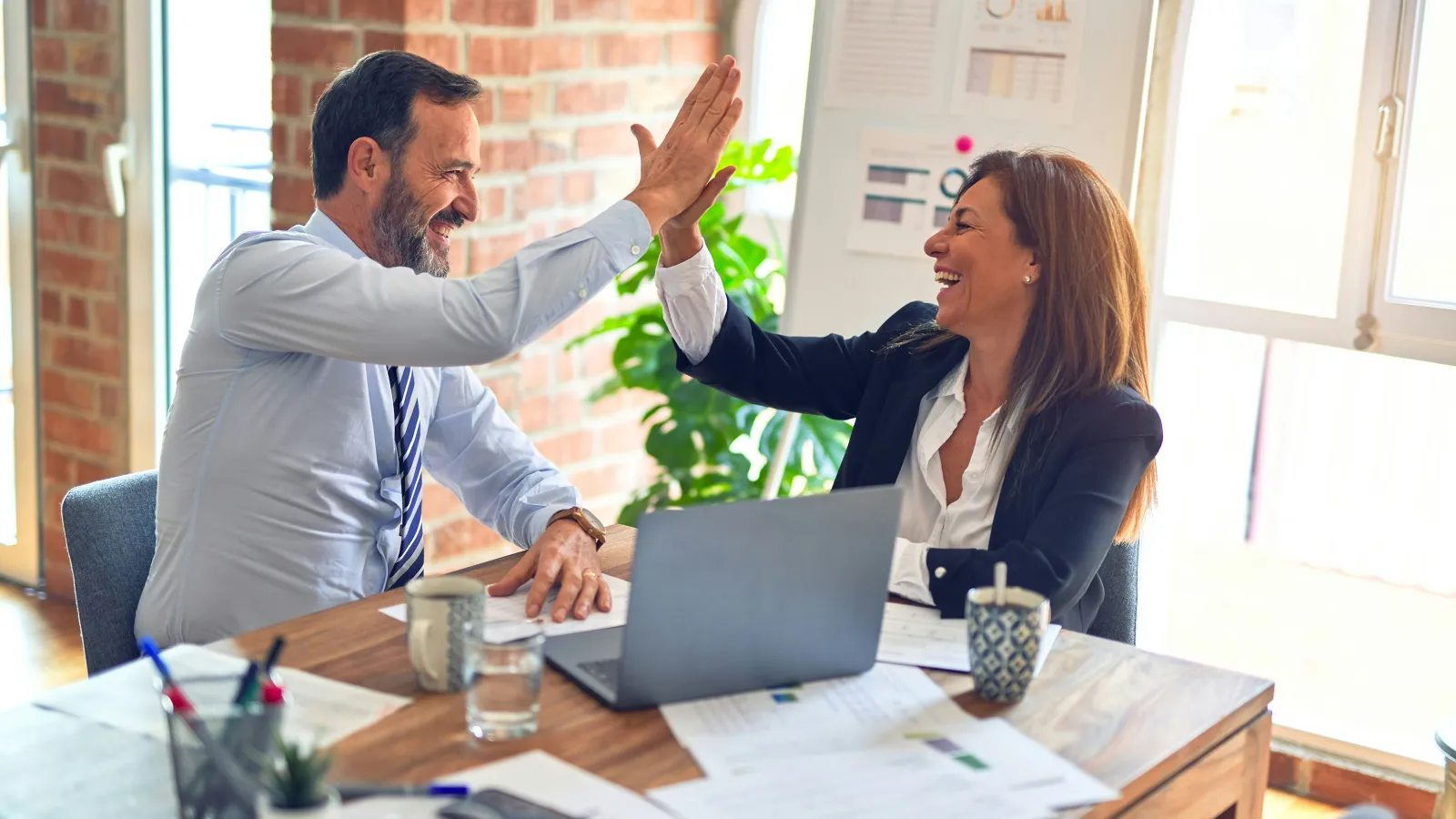 Business colleagues exchanging a high-five at a desk with laptop and documents in a bright office.