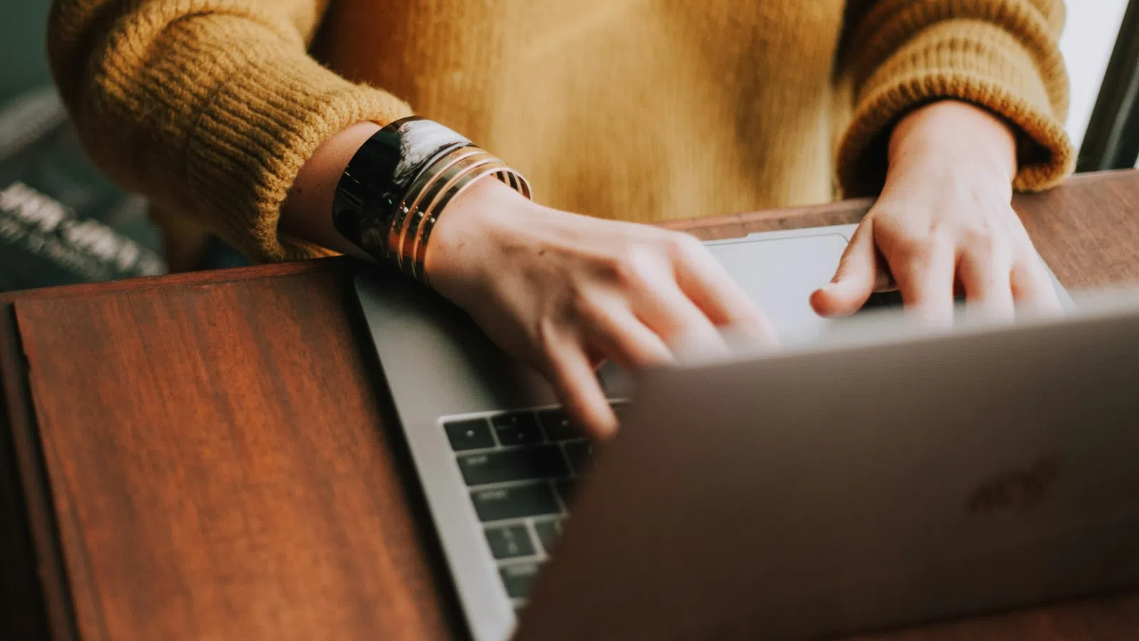 Person wearing a mustard sweater typing on a laptop at a wooden desk with a silver bracelet on the wrist