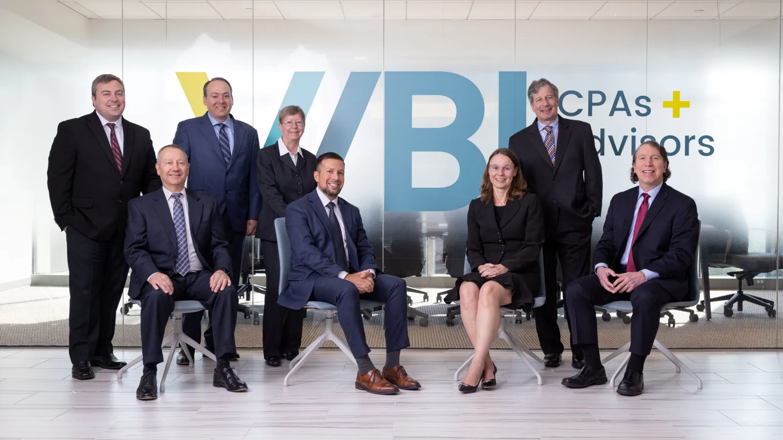 Professional group of eight business people posing in office with WBK CPAs and Advisors logo on glass wall behind.