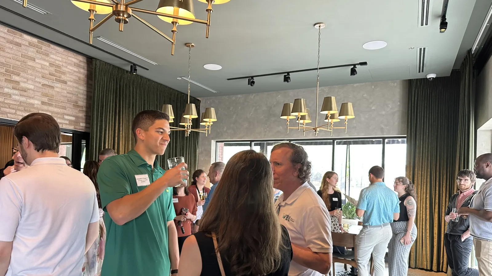 People networking and socializing in a modern, well-lit room with large windows and chandeliers.