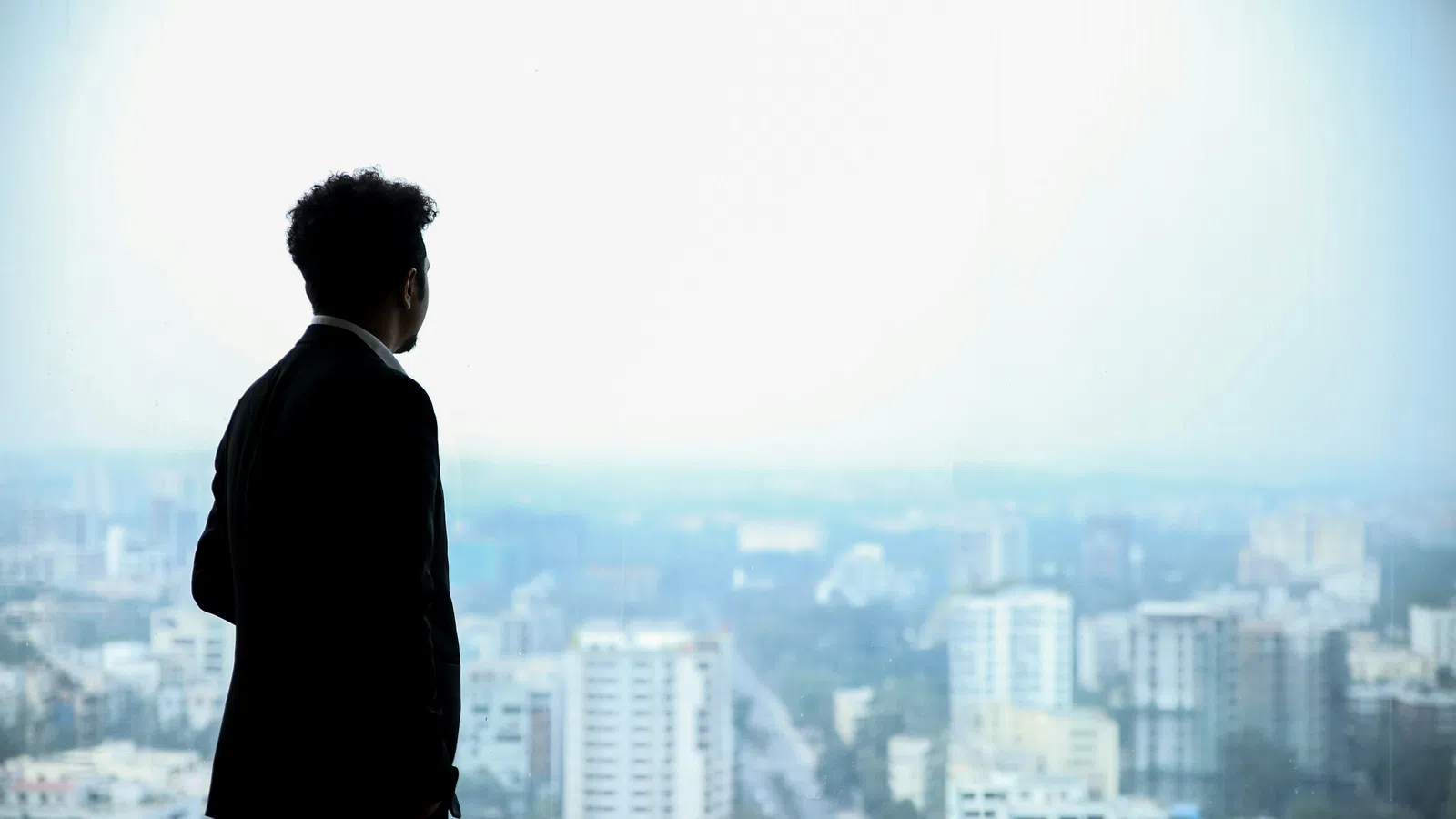 Man in suit standing by window overlooking city skyline with tall buildings on a foggy day.