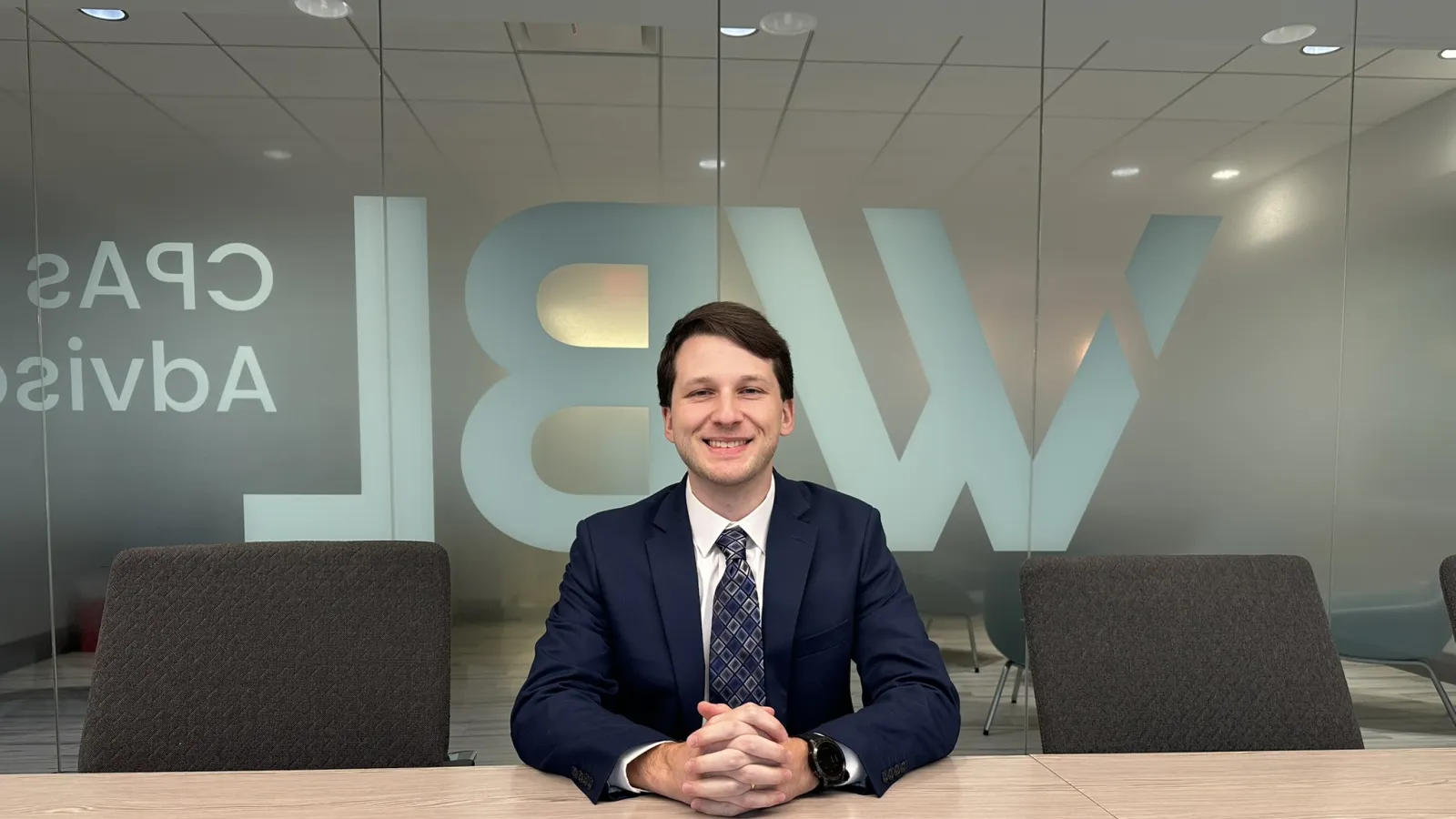 Young businessman in suit sitting at a conference table smiling in a modern office meeting room.