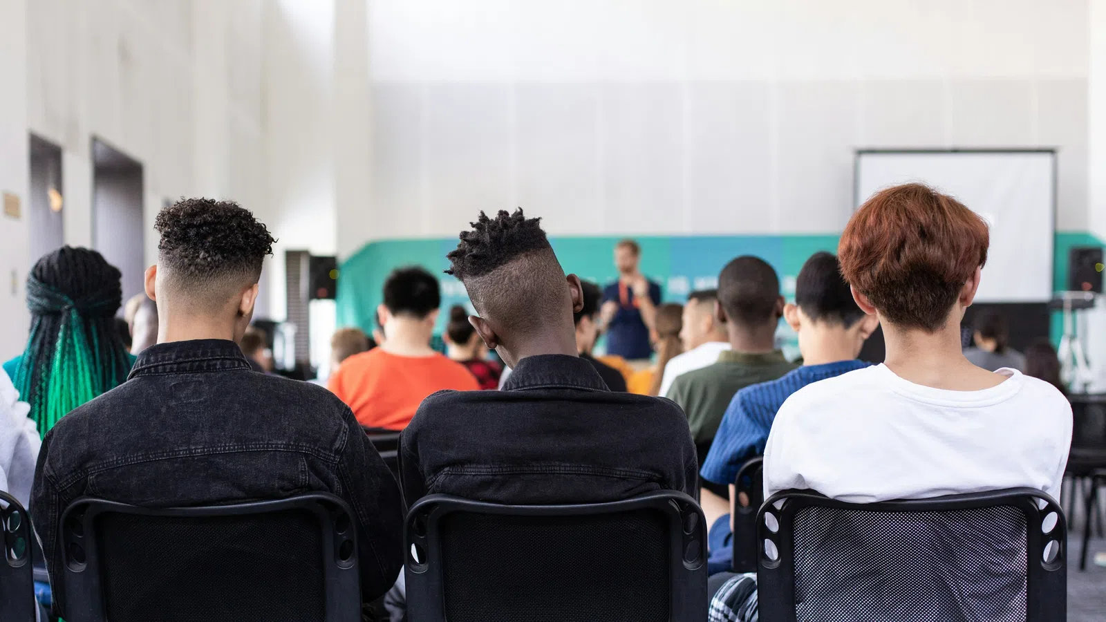 Diverse students seated in a classroom facing a speaker and projector screen during a lecture or presentation.