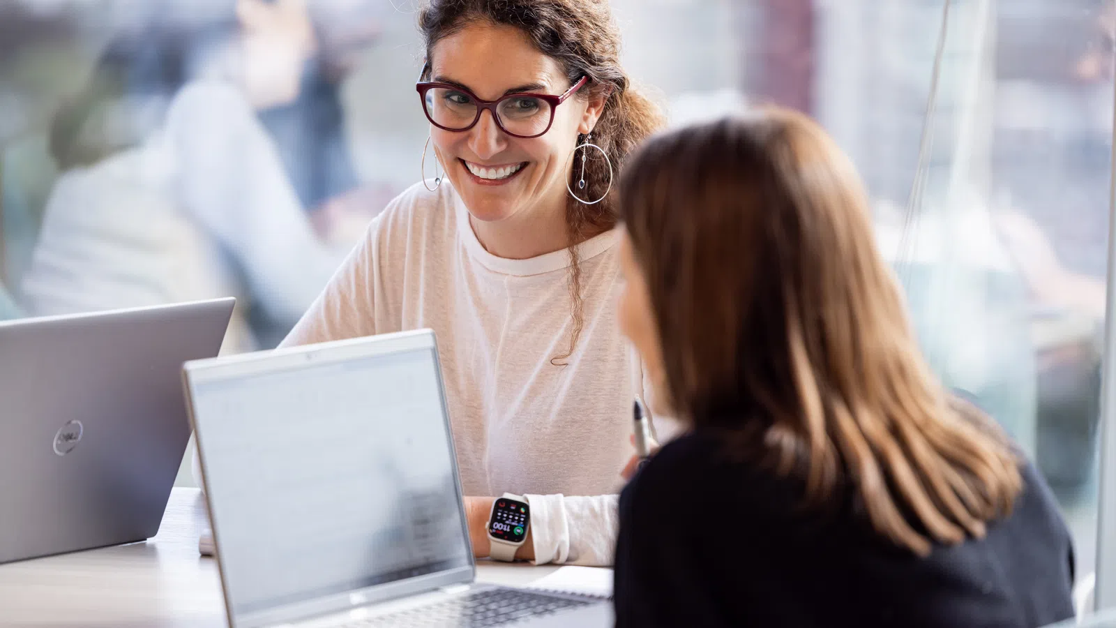 A smiling woman in glasses and a casual shirt converses with a friend at a table with laptops.