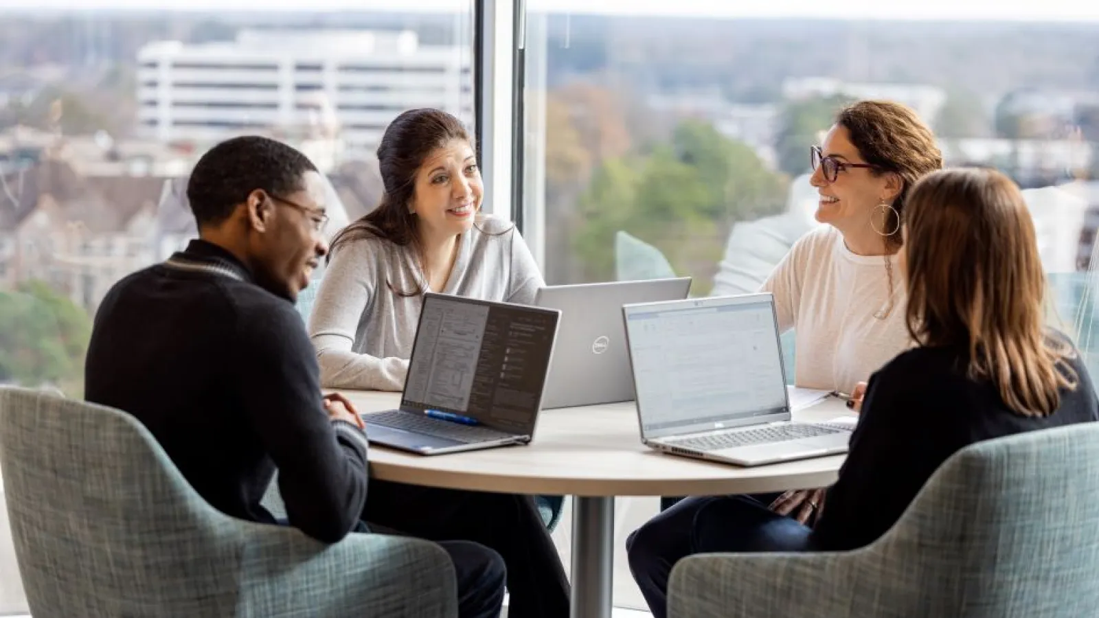 Four diverse professionals collaborating at a round table with laptops in a modern office overlooking the city.