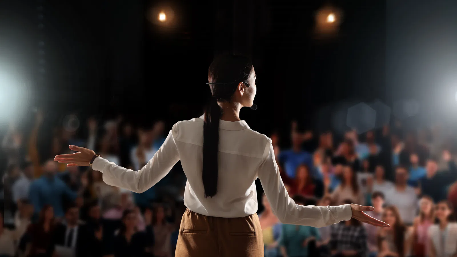 Woman speaker with microphone gesturing to an audience during a presentation in a dark auditorium