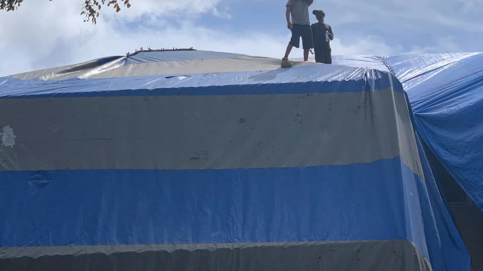 Two people standing on a large blue and gray tarp covering a roof under a cloudy sky