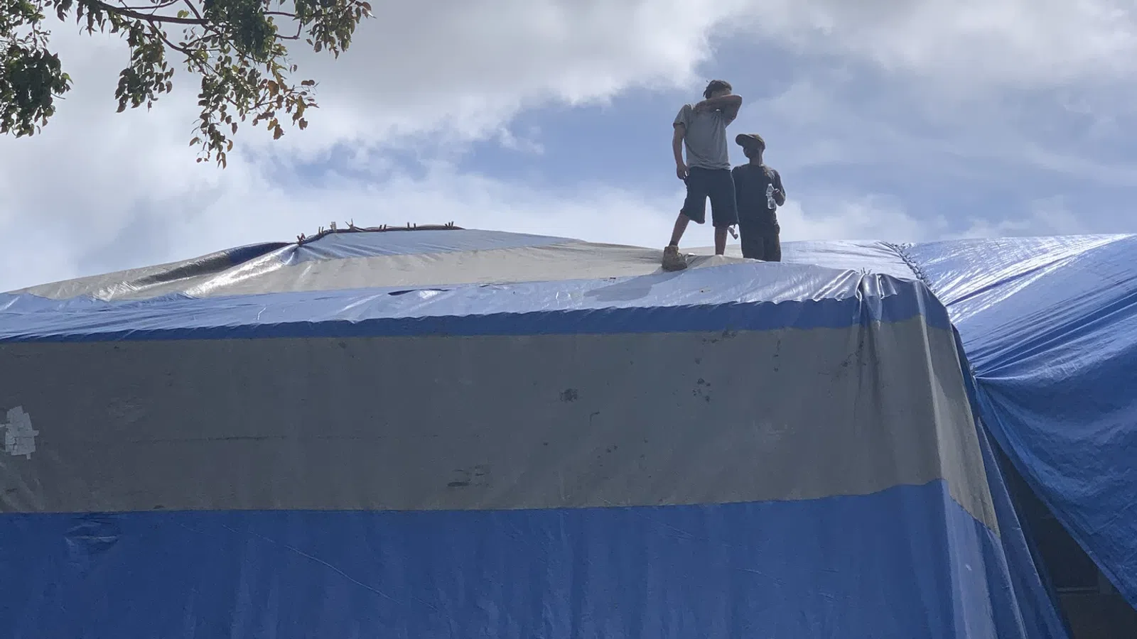Two people standing on top of a large blue and gray tarp-covered structure under cloudy sky near a tree.