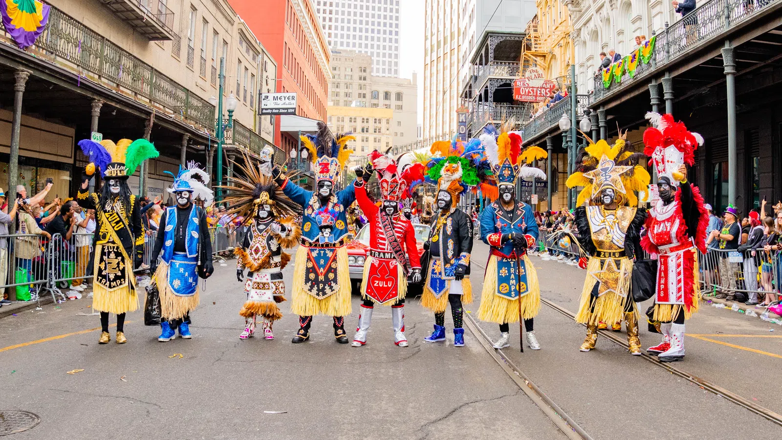 A colorful street parade with participants in feathered costumes and painted faces in an urban setting.