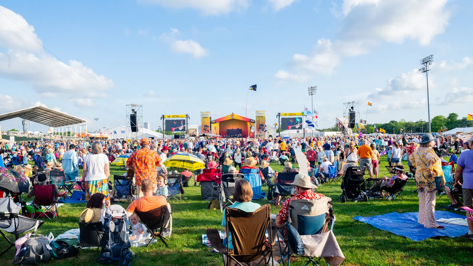 Large outdoor music festival crowd seated and standing on grass watching performances under blue sky with clouds.
