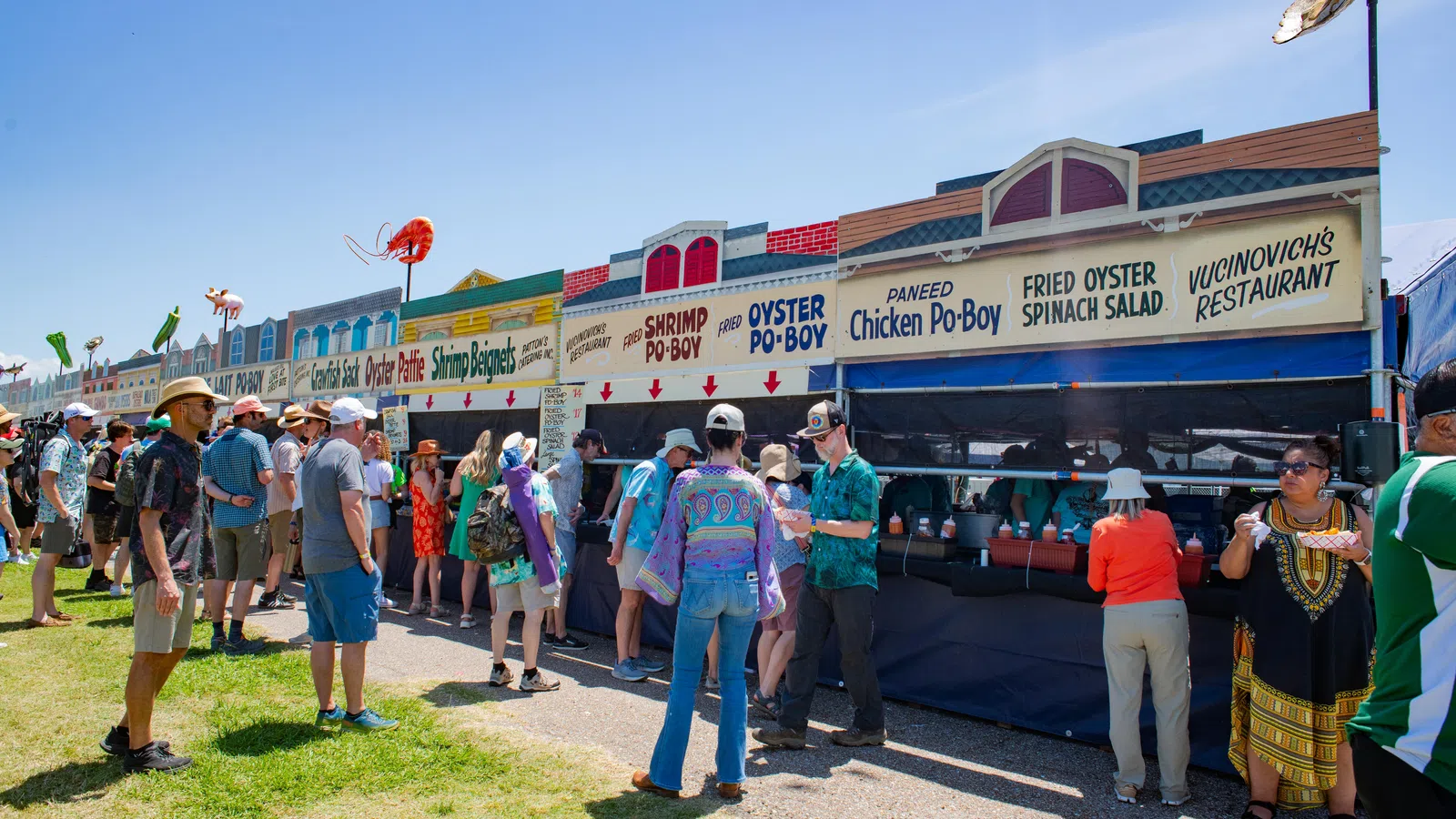 People lined up at outdoor food stalls serving seafood dishes like shrimp po'boy and fried oyster on a sunny day.