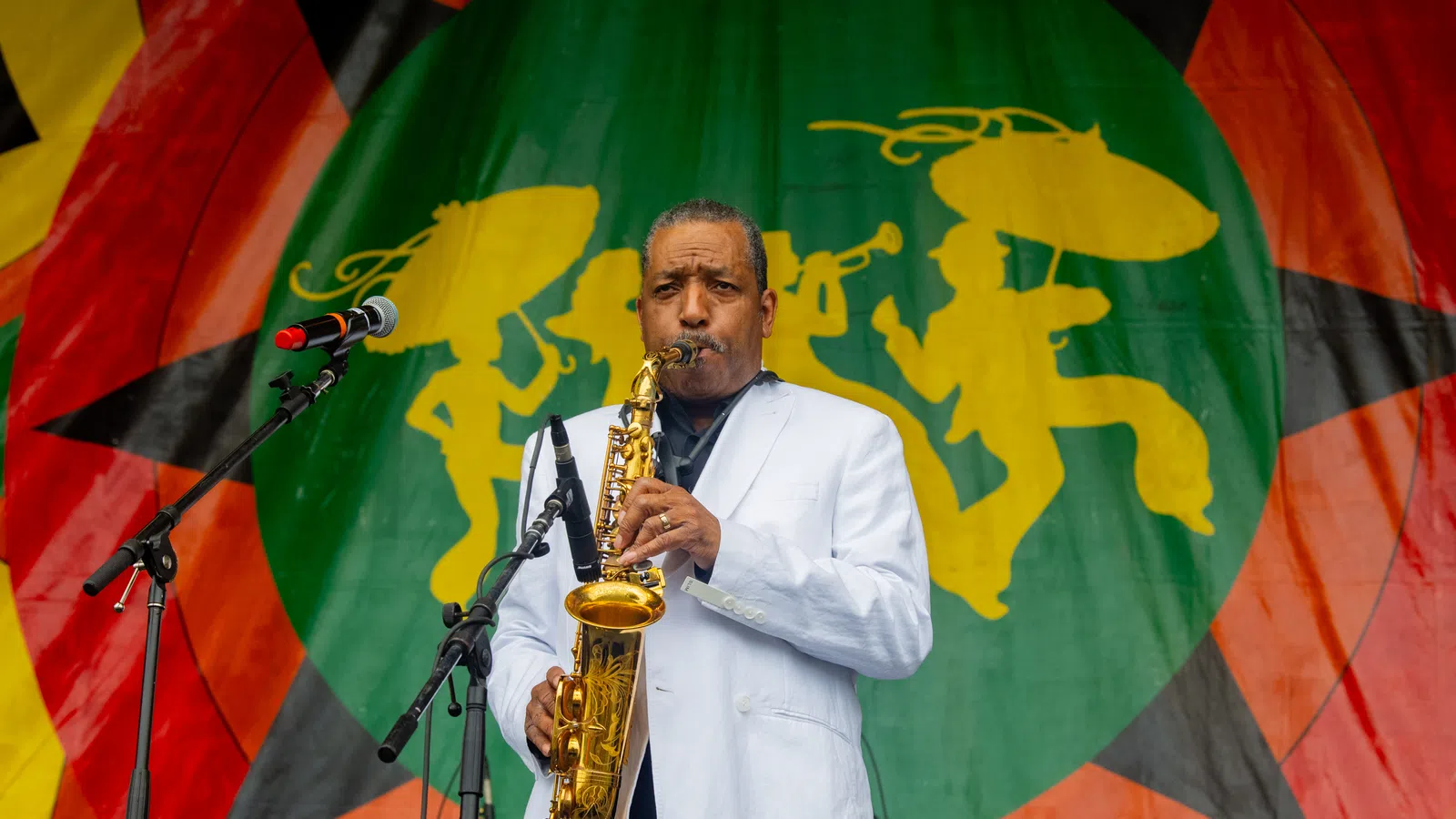 Saxophonist in white jacket performing on stage with colorful backdrop featuring silhouettes of musicians.