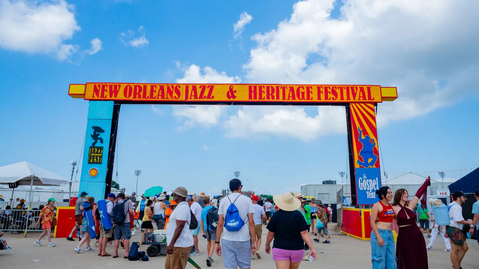 Crowd walking under New Orleans Jazz & Heritage Festival entrance arch on a sunny day with blue sky.