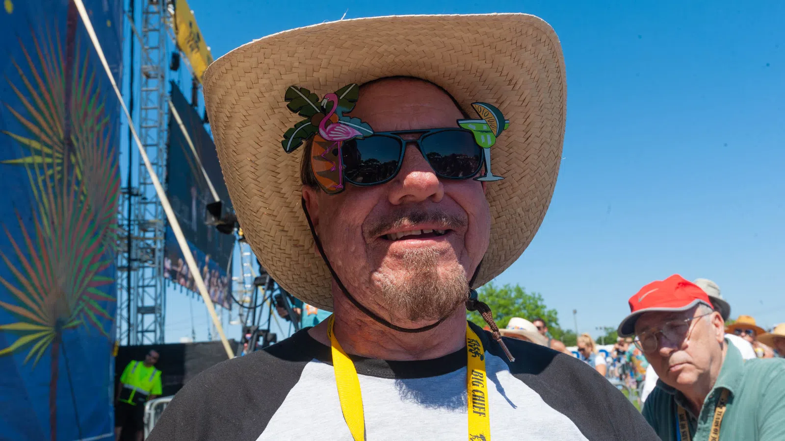 Smiling man wearing a straw hat and novelty sunglasses at an outdoor festival on a sunny day