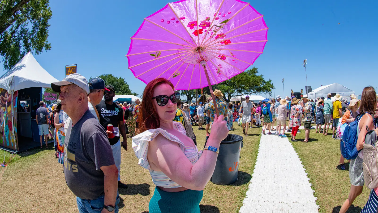 Woman holding a pink umbrella at a sunny outdoor festival with crowds and tents in the background