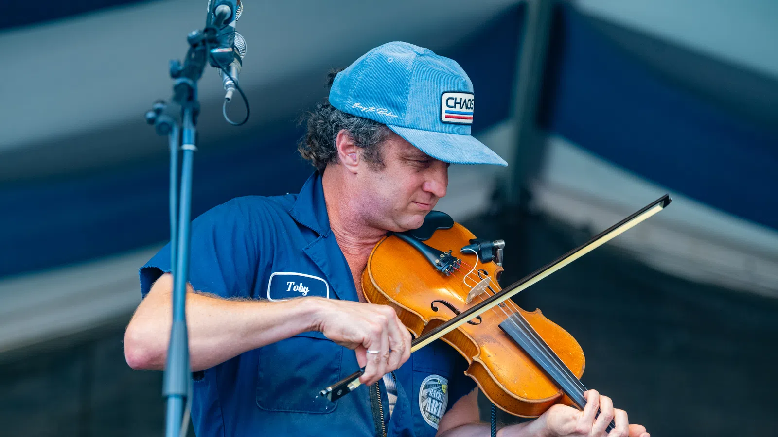 Musician wearing blue cap and shirt playing a violin on stage under a microphone.