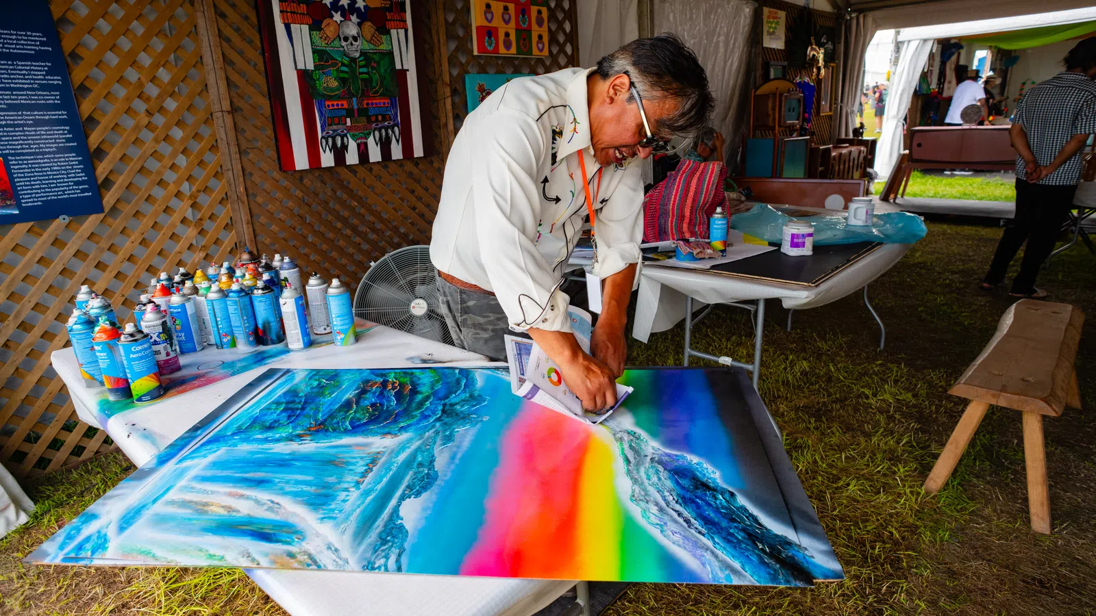 Artist creating a vibrant rainbow-themed painting indoors surrounded by spray paint cans and artwork.