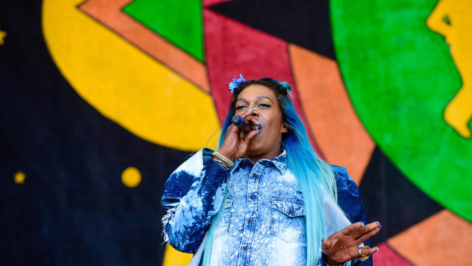 Female singer with blue ombre hair performing on stage with colorful abstract background art.