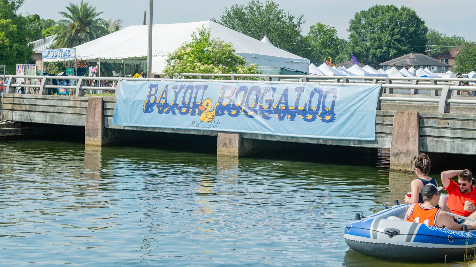 People in an inflatable boat on a bayou under a bridge with a Bayou Boogaloo festival banner.