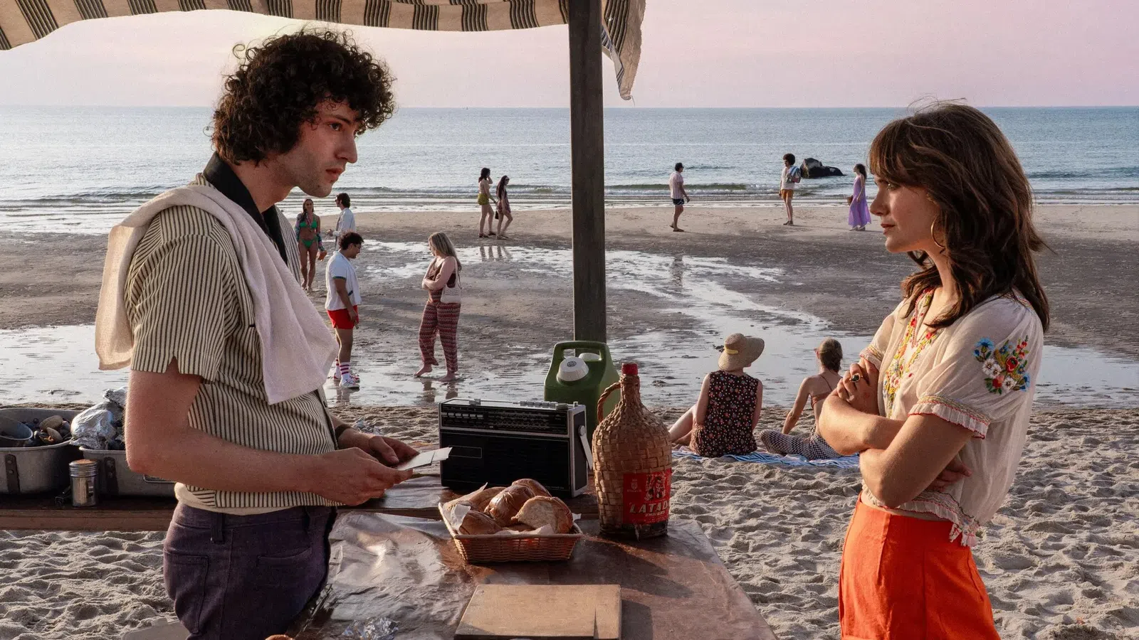 Young man and woman talking at a beachside stand with bread, drinks, and people walking along the shore at sunset.