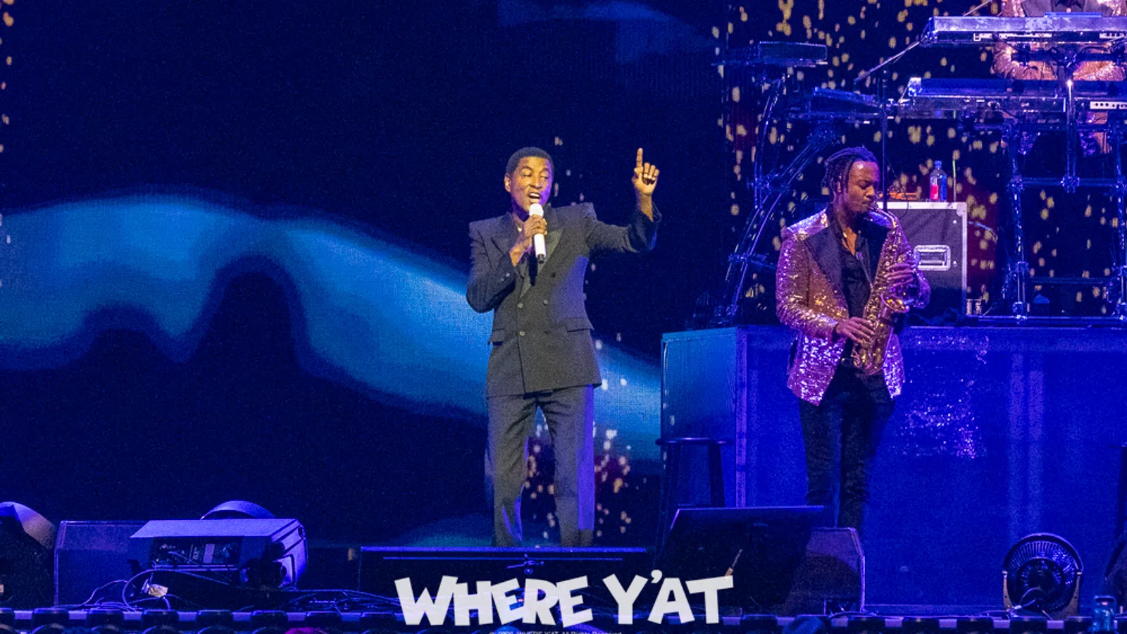 Singer performing on stage with saxophonist and DJ, vibrant lighting and audience visible in foreground