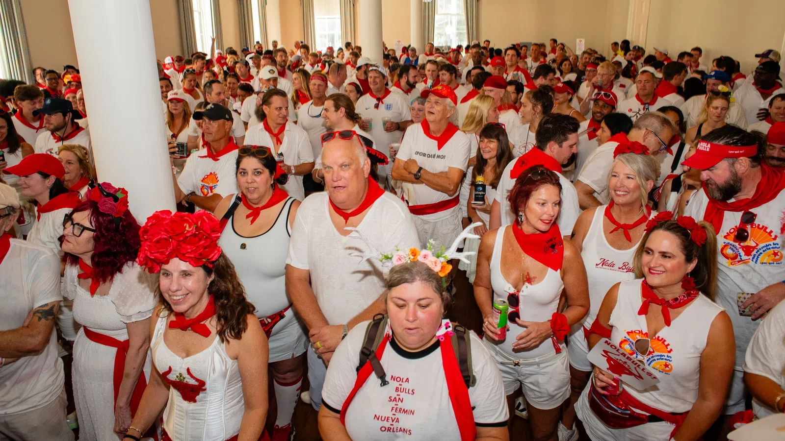 Large crowd dressed in white with red accessories gathers indoors celebrating a festive event.