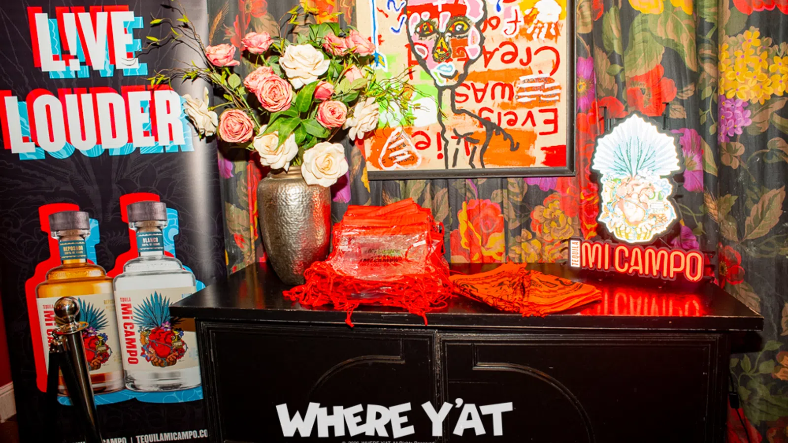 Colorful display with Live Louder banner, Mi Campo tequila bottles, floral vase, art, and Where Y'at signage on a black table.