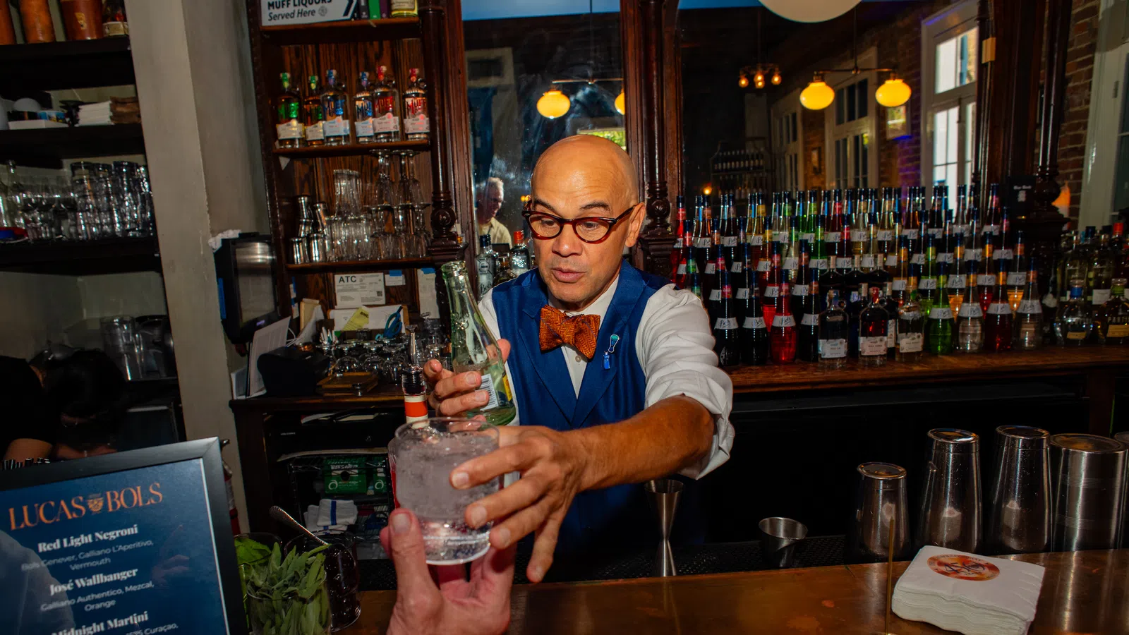 Bartender in a bow tie serving a cocktail at a wooden bar counter with bottles and glassware behind him
