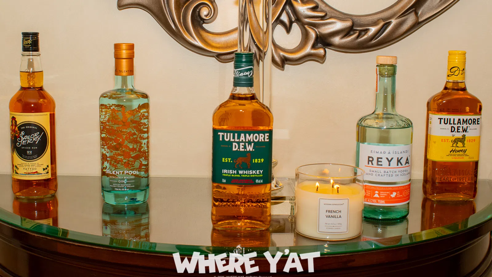 Five assorted liquor bottles and a lit French Vanilla candle displayed on a curved glass table with ornate metal wall decor.