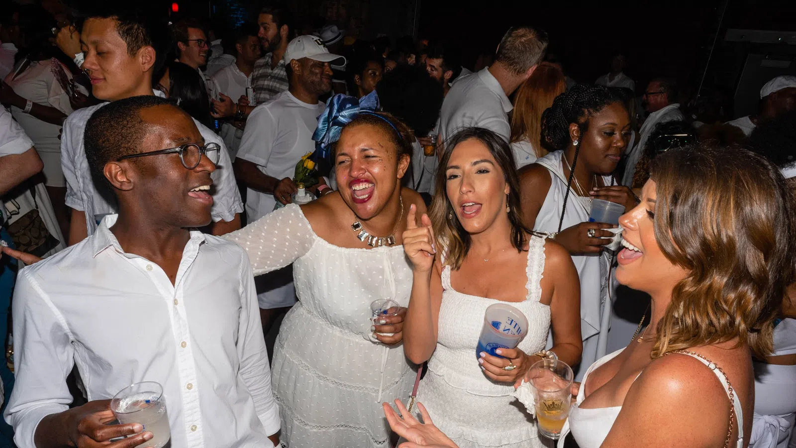 Group of friends in white outfits enjoying lively party with drinks and dancing at night event.
