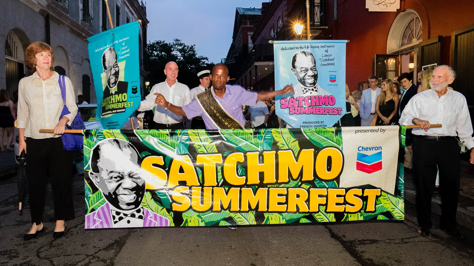 People holding a colorful Satchmo Summerfest banner during an evening parade in a city street.
