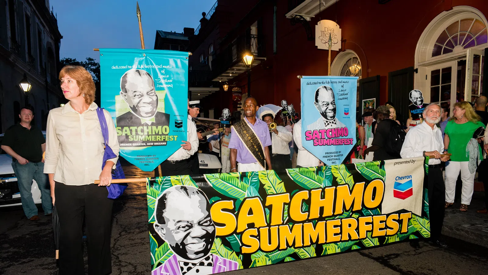 People holding colorful banners for Satchmo Summerfest during an evening street parade in New Orleans.