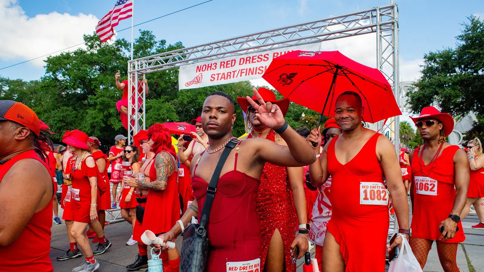 Participants dressed in red outfits posing at the NOH3 Red Dress Run start/finish line under a bright sky.