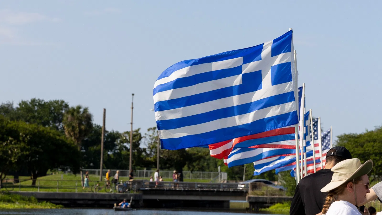 Greek flags waving alongside American flags by a riverside with people and greenery under a clear sky.