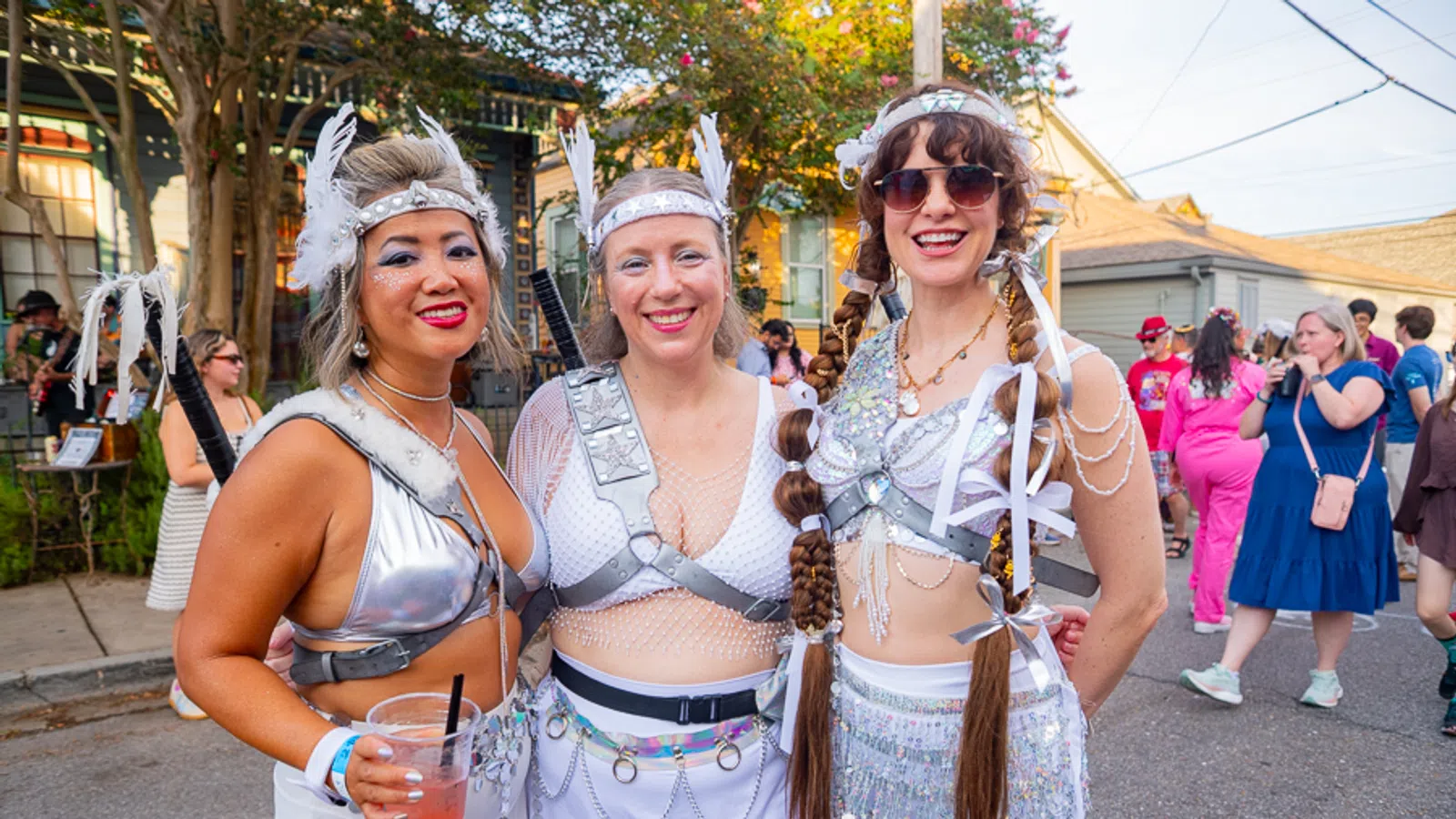 Three women in futuristic silver festival costumes posing outdoors at a sunny street event with a festive crowd.